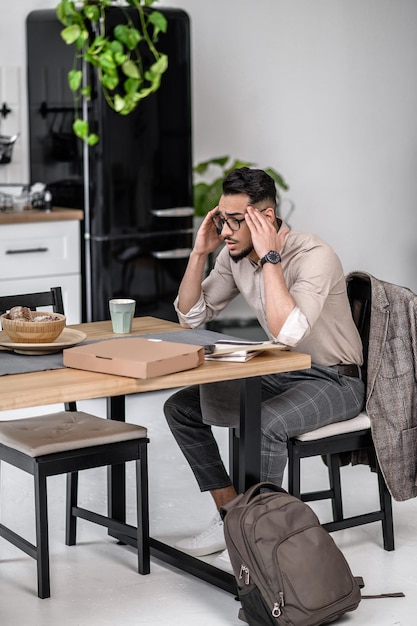 Tiredness. Young bearded man in glasses touching his head sitting tired at table with correspondence at home