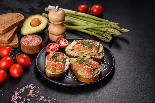 Toast sandwich with butter avocado and salmon decorated with arugula and sesame seeds on a black stone background