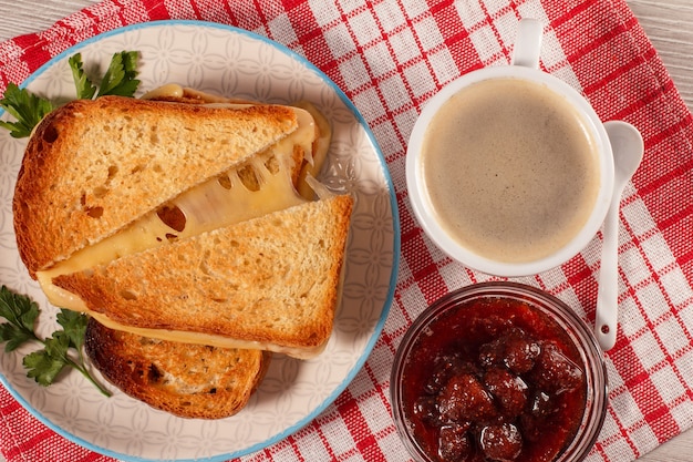 Toasted slices of bread with cheese and green parsley on white plate, cup of coffee, glass bowl with strawberry jam and spoon with red kitchen napkin. Top view. Good food for breakfast