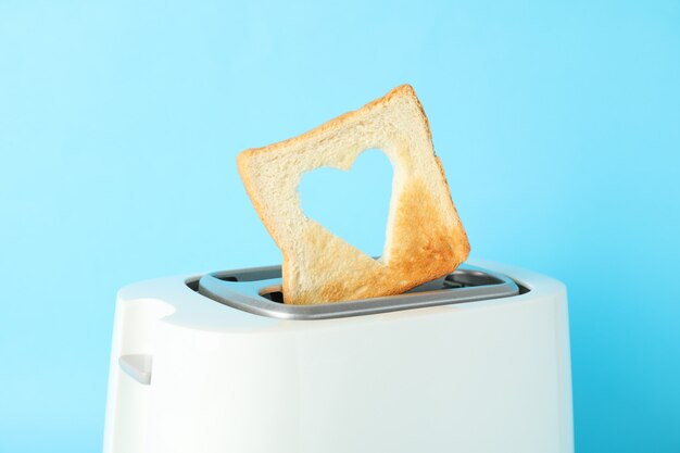 Toaster and bread slice with a shape of heart on blue background, close up