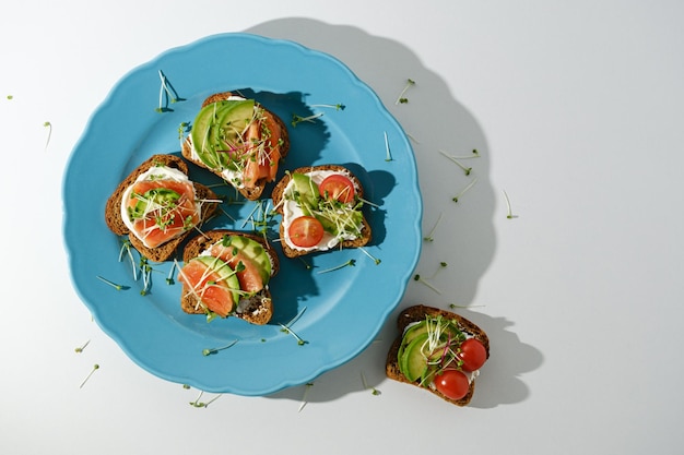 Toasts with salmon cottage cheese avocado microgreens cherry tomatoes in blue plate on white background open fish sandwiches top view