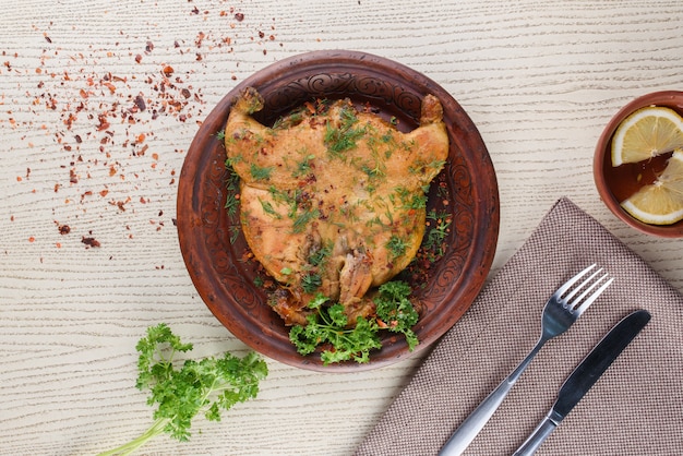 Tobacco chicken decorated with herbs on a clay plate on a white wooden table. Top view.