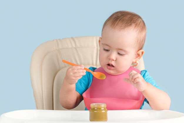 toddler aged 12-23 months sits on a feeding chair and learns to eat broccoli puree from a glass jar of baby food with a spoon on his own