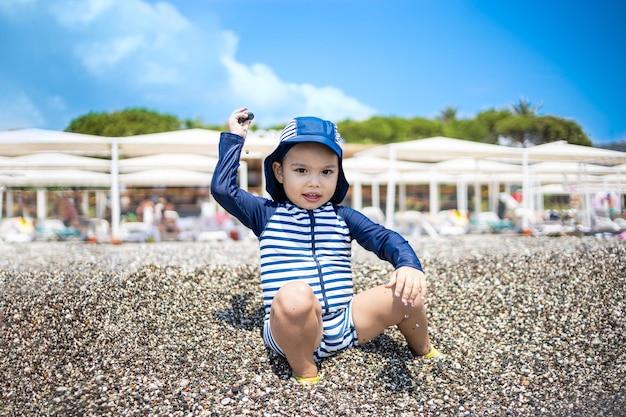 Toddler boy in a swimming suit plays with pebbles on the seashore in a hotel