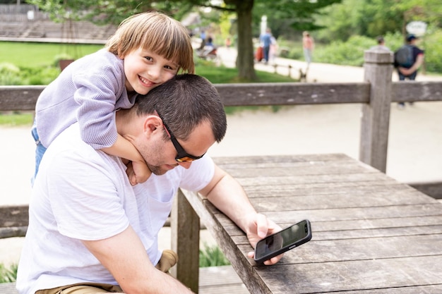 Toddler boy tightly hugs his dad looking at the smartphone in the park in summer Child disturbing his dad while using internet and working