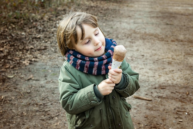 Toddler boy wearing warm jacket and scarf eating chocolate ice cream in waffle cone with pleasue outdoor in cold weather