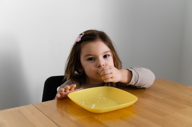 Toddler girl eating bread or pie at home with her hands. Hungry kid. Unhealthy diet. Bad table