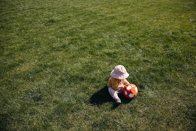 Toddler playing with a red football ball on a meadow with green grass on a spring day