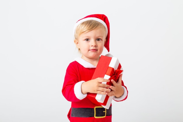 A toddler in a Santa costume stands on a white background next to gift boxes for Christmas. Christmas concept, text space