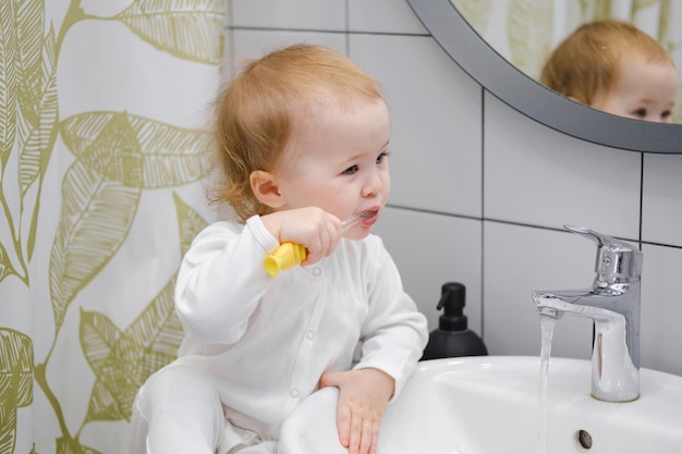 A toddler sitting on the washbasin cabinet and brushing teeth showing independence