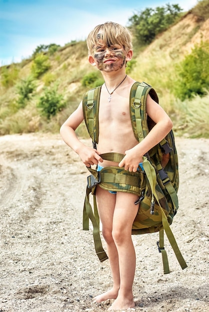 Toddler with dirty painted face and camouflage backpack stands on beach against hills