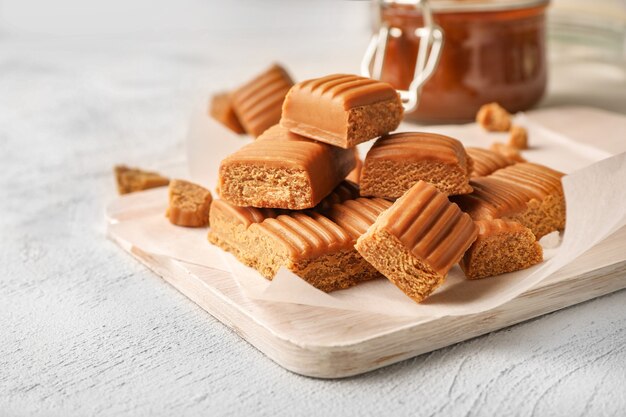 Toffee candies on cutting board with melted caramel in glass jar nearby on white