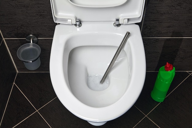 A toilet bowl with a toilet brush and cleaner and disinfectant on a dark ceramic tile background