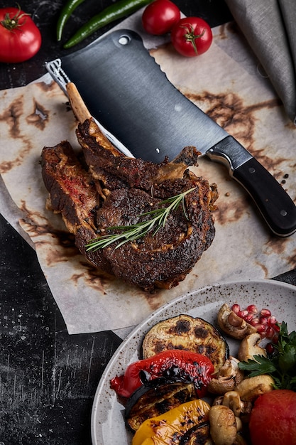 Tomahawk steak with vegetables and a knife on the table. Grilled meat with grilled vegetables and fresh vegetables on the table.