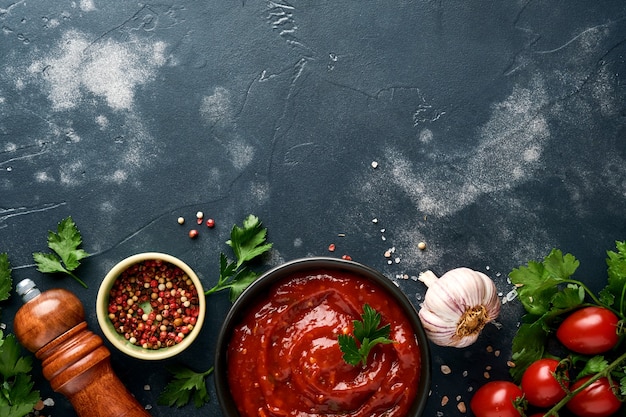 Tomato ketchup spicy sauce in a black bowl with spices and fresh tomatoes on a dark slate, stone or concrete background. Top view with copy space.