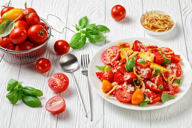 Tomato salad with crispy fried onion, quinoa and fresh basil on a white plate on a wooden table with ingredients, close-up, landscape view from above