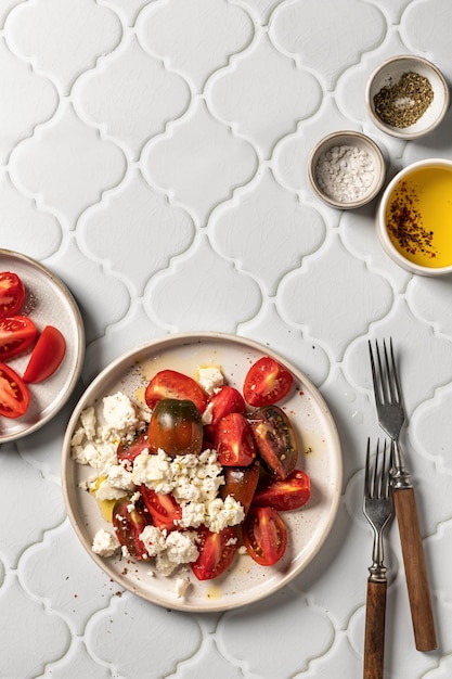 Tomato salad with feta cheese and herbs served with cutlery on white tile background top view