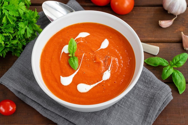 Tomato soup puree in a bowl with cream and fresh basil leaves on a dark wooden background Top view