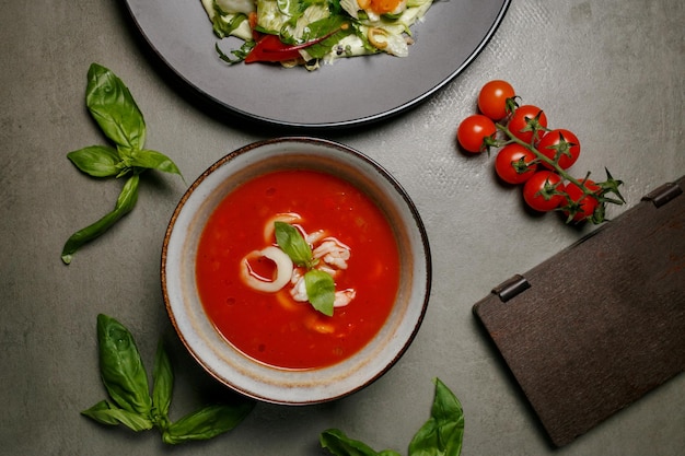 tomato soup with squid in a plate on a gray background, next to a wooden menu and tomatoes, top view