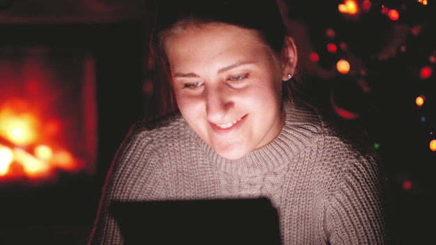 Toned footage of smiling young woman watching video on tablet at night in living room wiht burning fireplace