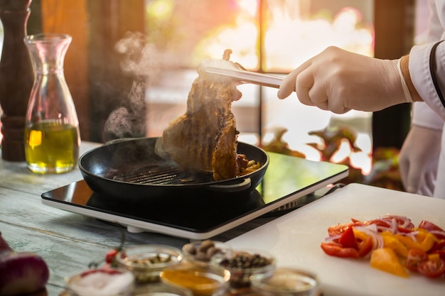 Tongs hold fried meat. Pieces of tomato and onion. Cook is preparing pork. Your lunch is almost ready.