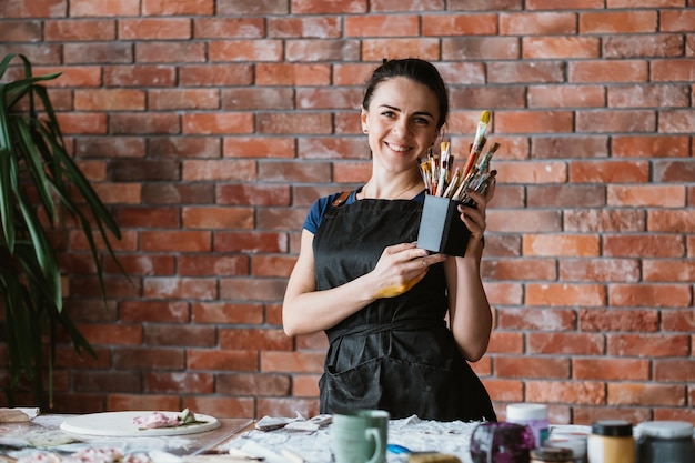 Tools for art talent. Woman painter workplace. Beautiful smiling female in apron posing with paintbrushes.