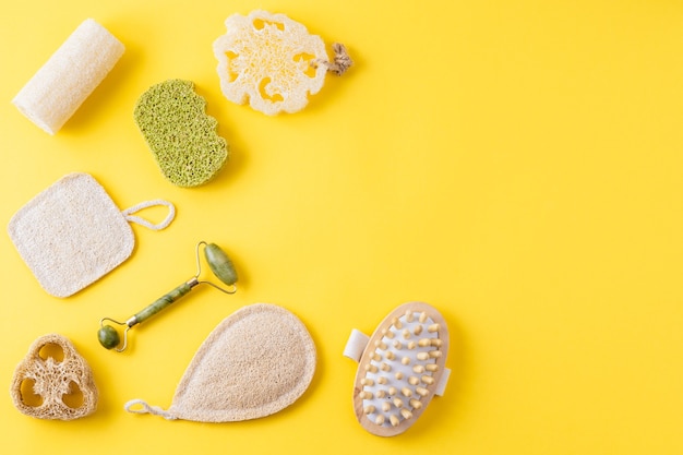 Tools for beauty routine from ecological material. Zero waste bathroom. Jade face roller, anti cellulite massager, loofah and bamboo sponges on yellow surface. Copy space, flat lay