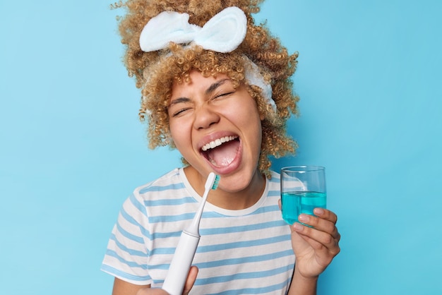 Tooth care concept Positive young woman with curly combed hair holds electric toothbrush and glass of mouthwash sings and has fun during morning routine wears headband casual striped t shirt