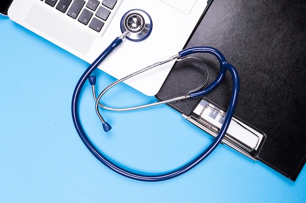 Top close up view of stethoscope next to a laptop and black folder on blue background