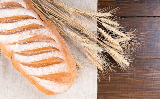 Top down view of large white bread loaf with a light coating of flour on paper next to stalks of dry wheat over dark wooden table