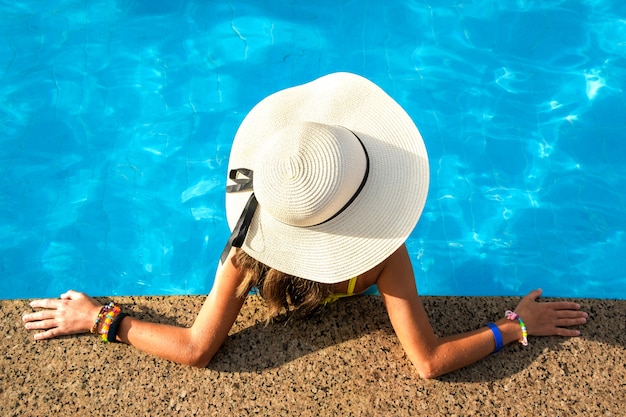 Top down view of young woman wearing yellow straw hat resting in swimming pool with clear blue water on summer sunny day.