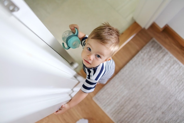 Top view of adorable blond little boy with beautiful blue eyes holding his bottle with water and looking at camera.