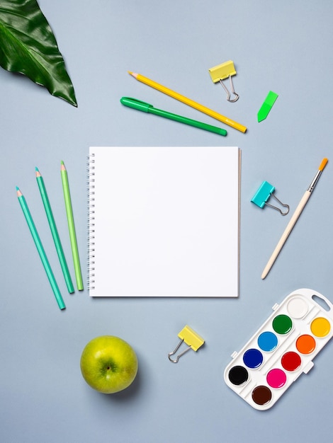 Top view arrangement with notepad with white sheets and pens pencils paper clips and other office supplies on blue background