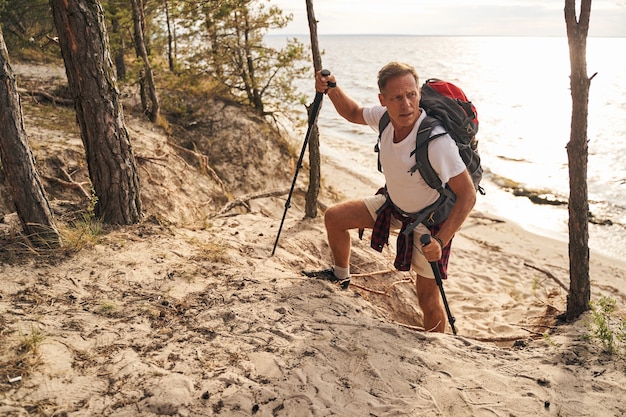 Top view of athletic mature male going up hill above sea shore while going nordic walking