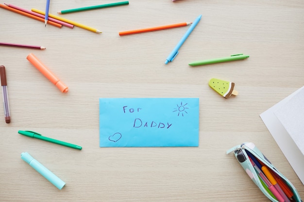 Top view background image of letter for dad with handmade gift on Fathers day set on table with colored crayons, copy space