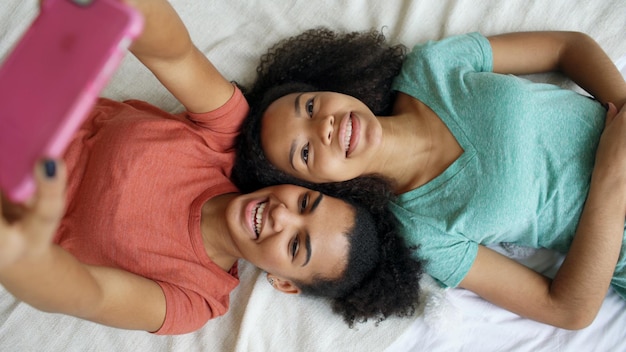 Top view of beautiful mixed race funny girlfriends making selfie portrait on bed in bedroom at home