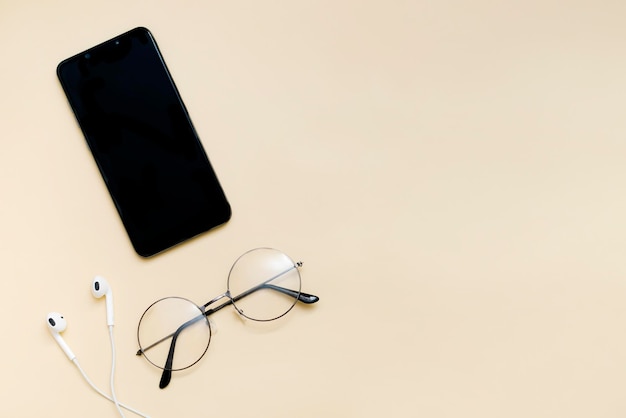 Top view of black smartphone with blank screen and wire headphones glasses goggles for reading mobile phone as music gadgets on beige background top view copy space mock up template logo design