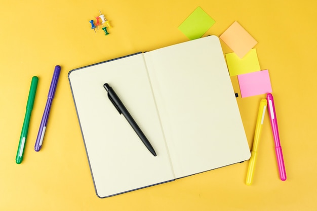 Top view of blank notebook and school supplies like colored markers, sticker and clipers on yellow background, space for text.