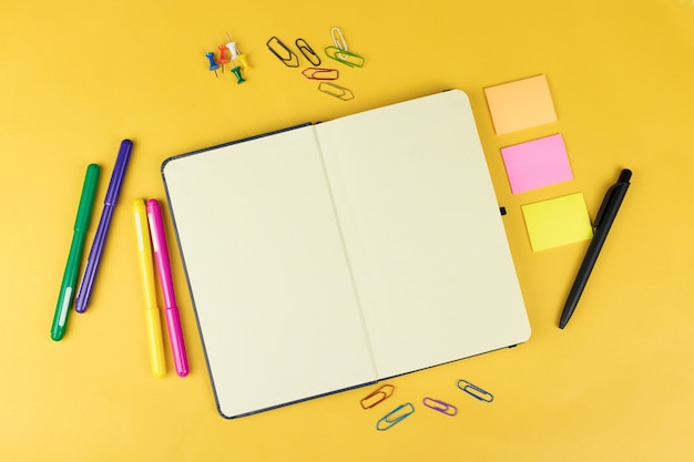 Top view of blank notebook and school supplies like colored markers, sticker and clipers on yellow background, space for text.