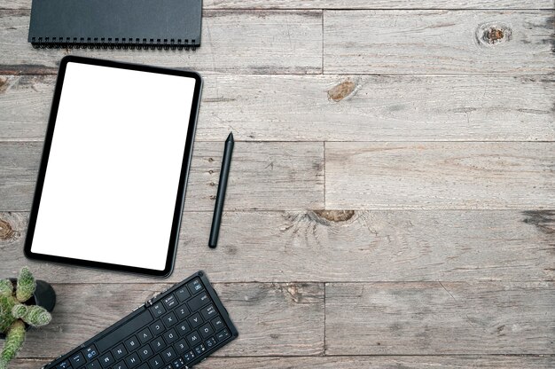 Top view blank screen tablet, pen, keyboard and notebook on wooden table background.