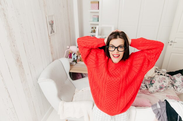 Photo top view of blue eyed, brunette young female in casual red sweater, wears rectangular spectacles, stands in her spacious room at home