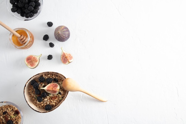 Top view of the bowl with homemade granola or muesli with figs and blackberries a jar of honey white background a copy of the space