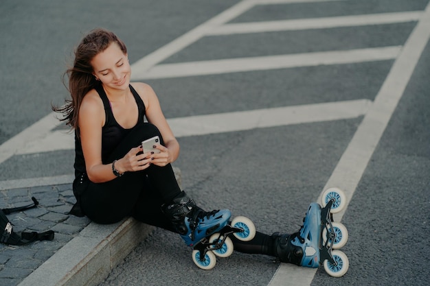 Top view of brunette active young woman uses smartphone for online communication poses on asphalt dressed in black active wear enjoys favorite hobby and free time Extreme sport and rest concept