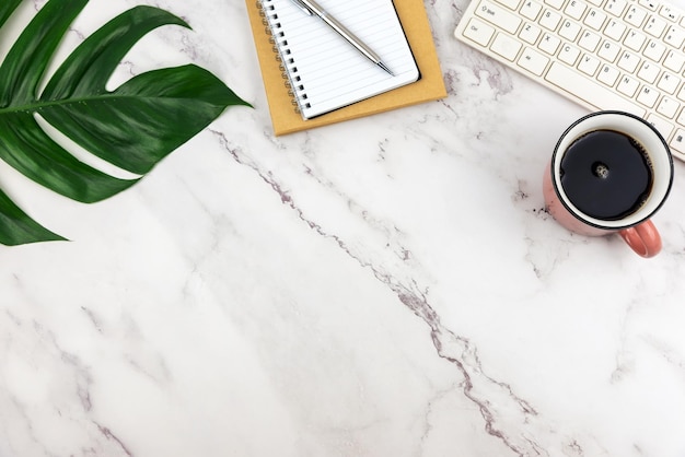 Top view of business desk table with tropical leaf and cup of coffee white marble pattern background