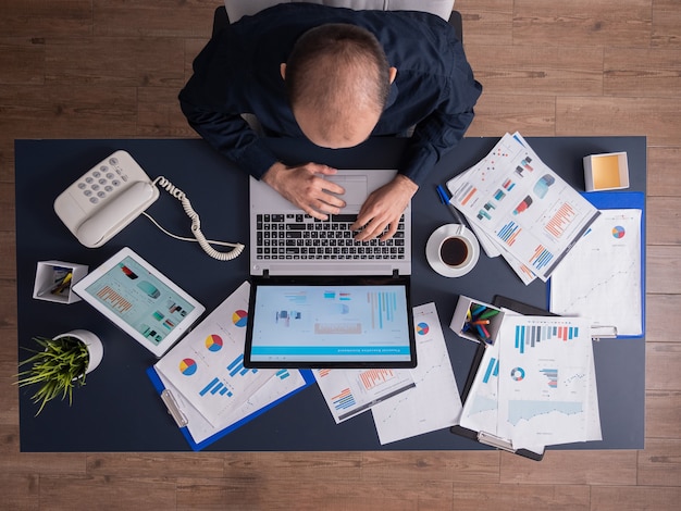 Top view of business man in corporate office sitting at desk, typing on laptop, working on financial statistics and business strategy. Entrepreneur using touchepad scrolling through documents.