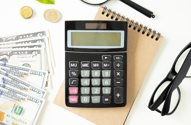 Top view of calculator, notepad, cups with coffee, coins and green plant on gray table.