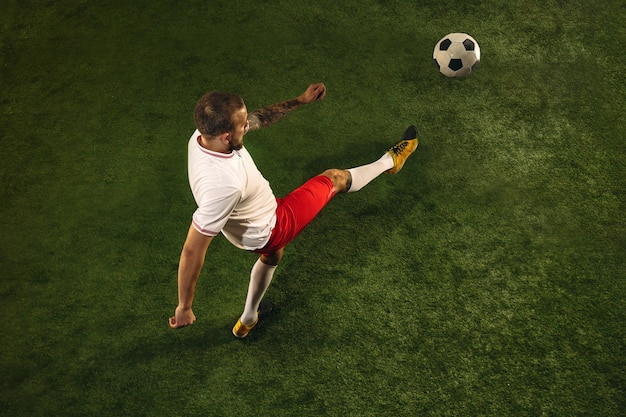 Top view of caucasian football or soccer player on green background of grass. Young male sportive model training, practicing. Kicking ball, attacking, catching. Concept of sport, competition, winning.