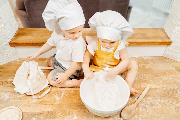 Top view of charming curious children brother and sister in a suit of cooks are sitting on the kitchen table and preparing dough for pancakes. The concept of teaching children to work and cooking