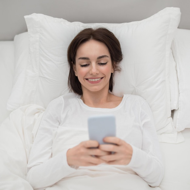 Top view of cheerful young woman using phone in bed