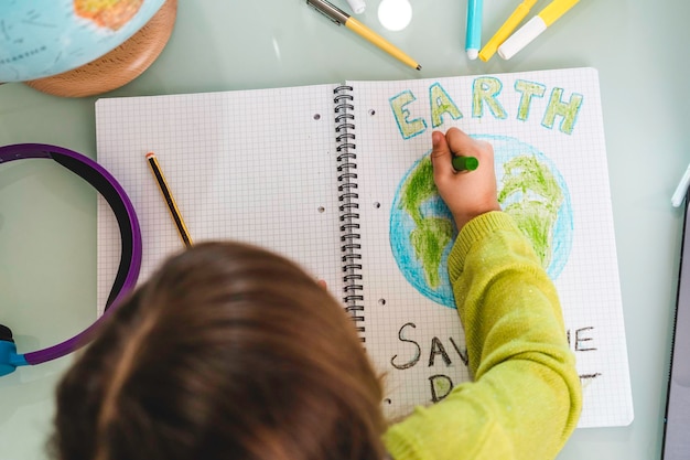 Top view of child girl draws planet earth with wax colors on school notebook for Earth day Little activist girl writes the message Save the Planet Protection of environment global warming